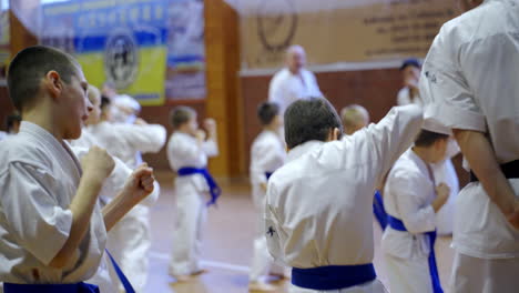 athletes and their coaches in karate having training in a large gym. kids in white kimonos make moves with hands. blurred backdrop.
