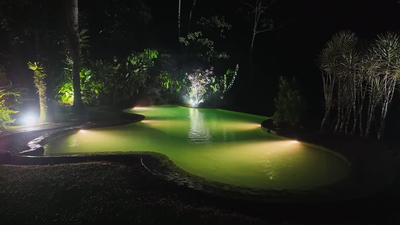 piscina iluminada vacía con mini cascada en un jardín tropical por la noche en costa rica
