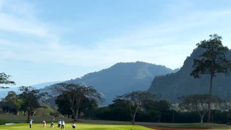 golfers walk across a vibrant green course with stunning mountain scenery in the background.