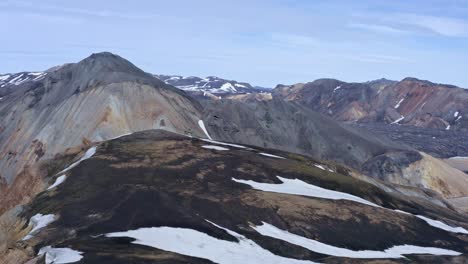 Vista-Frontal-De-Drones-Sobre-Las-Montañas-De-Riolita-De-Landmannalaugar,-Frente-A-Las-Montañas-Azules-De-Bláhnúkur,-Que-Muestran-Paisajes-De-Verano-Con-Restos-De-Nieve.