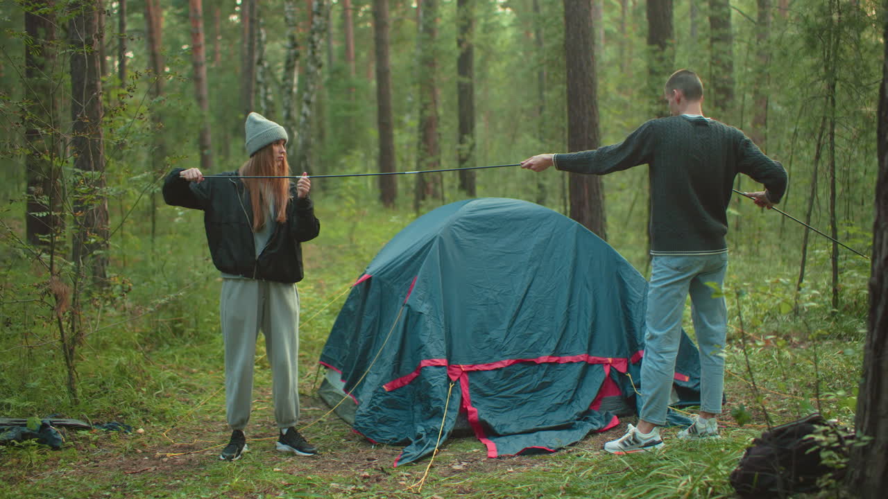 Couple standing in forest adjusting flexible tent pole with pitched tent behind them, surrounded by trees and grassy terrain, collaborating on outdoor camping setup under natural daylight