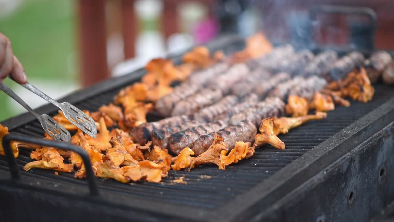 Man preparing sausage and mushrooms on the grill using tongs. Slow motion shot. Close up
