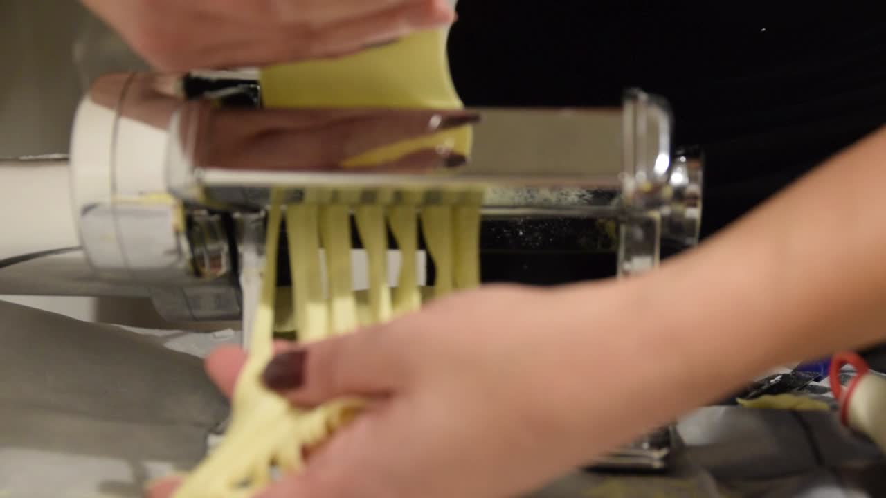 Mani che raccolgono tagliatelle fresche da una tradizionale macchina per la pasta in ferro, vista frontale.