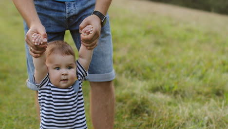 el primer plano de un bebé que da sus primeros pasos mientras camina descalzo sobre la hierba verde y sostiene las manos de su padre