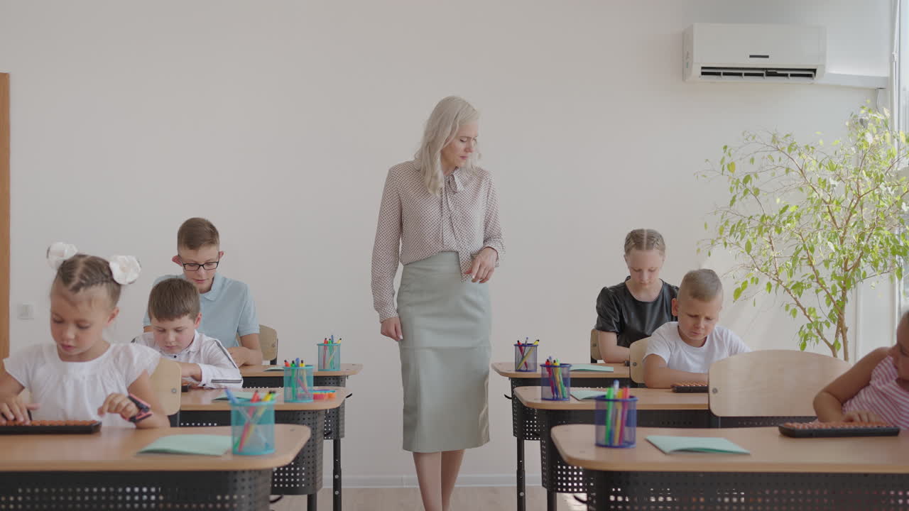 In Elementary School Class: Enthusiastic Teacher walks between Rows of Bright Diverse Children Explains Lesson. Group of Smart Multiethnic Kids Learning New Stuff. Side View Tracking Shot
