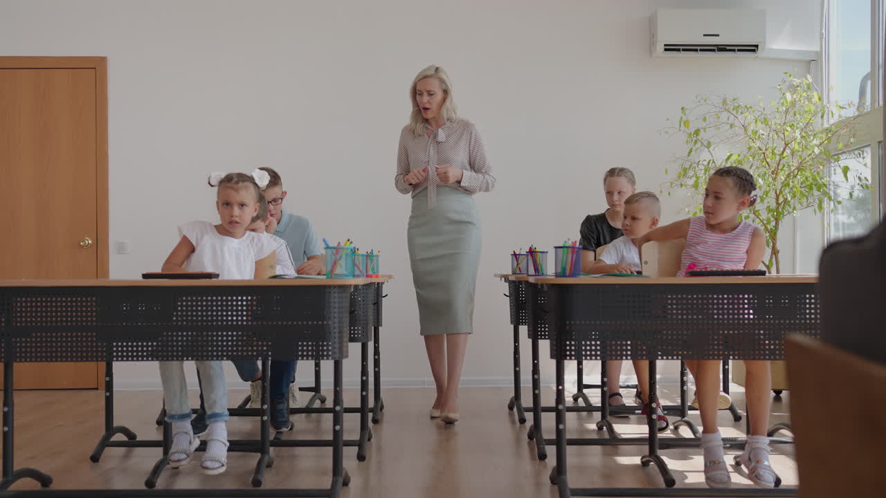In Elementary School Class: Enthusiastic Teacher walks between Rows of Bright Diverse Children Explains Lesson. Group of Smart Multiethnic Kids Learning New Stuff. Side View Tracking Shot
