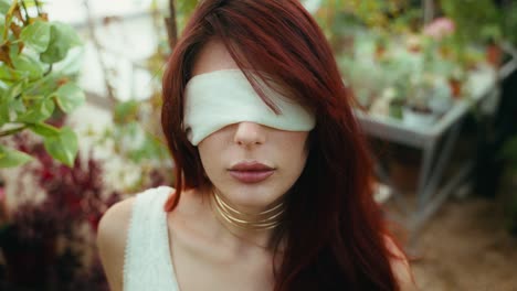 close-up-high-angle-shot-of-young-woman-in-white-dress-and-blindfolded-looking-at-camera-in-greenhouse-plant