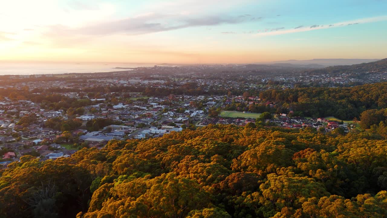 Aerial establishing over forested hills to reveal Illawarra suburbs framed by distant coastline at holden hour