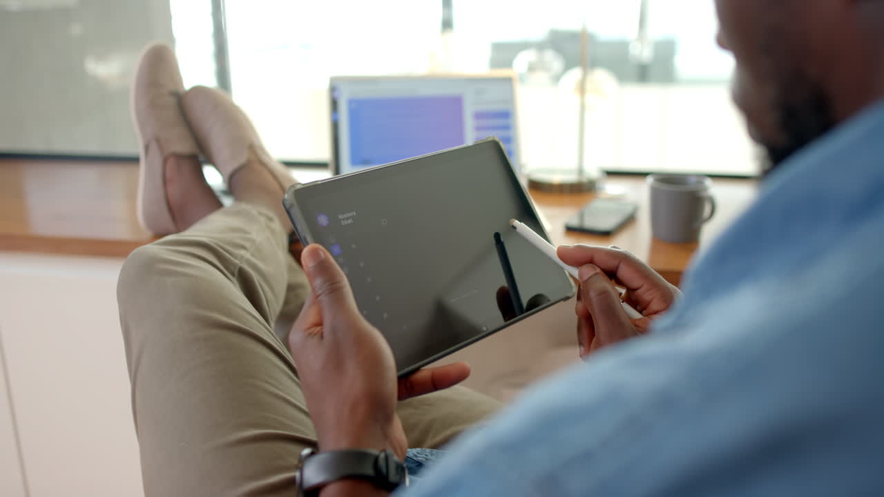 Using stylus on tablet, African American man relaxing with feet up at home office