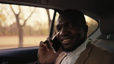 Portrait-of-a-happy-Black-skinned-businessman-in-a-brown-jacket-who-communicates-on-the-phone-while-driving-in-the-passenger-seat-in-a-modern-car-outside-the-city-during-a-business-trip