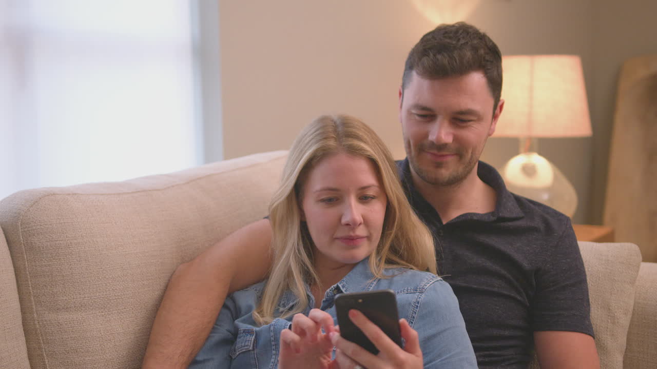 Evening shot of couple relaxing on sofa at home looking at mobile phone - shot in slow motion