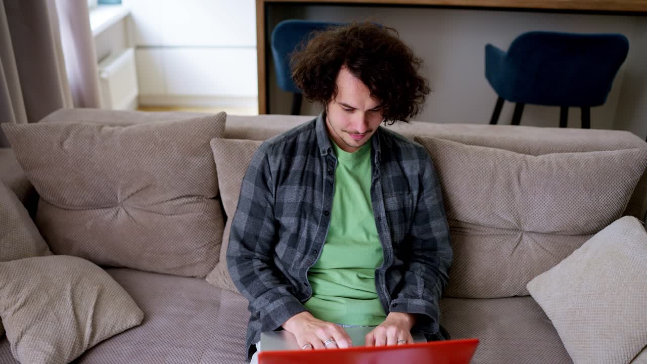 Happy brunette guy with curly hair in a gray checkered shirt sits on a light brown shirt at a laptop in a modern apartment