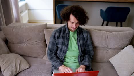 Happy-brunette-guy-with-curly-hair-in-a-gray-checkered-shirt-sits-on-a-light-brown-shirt-at-a-laptop-in-a-modern-apartment