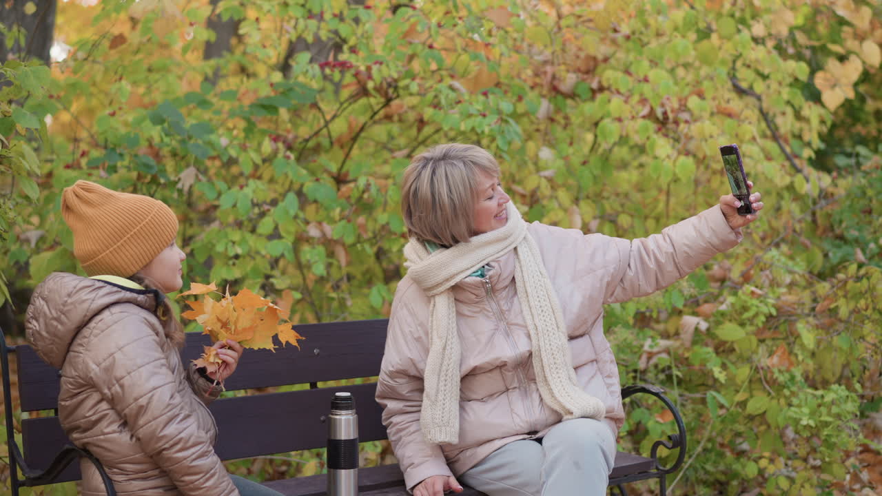 Middle aged woman in scarf and winter coat smiles as she takes selfie with smartphone while sitting on bench beside young girl holding yellow autumn leaves in peaceful park full of colorful foliage
