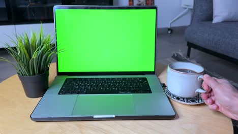 close-up of a hand holding a coffee cup beside a laptop with a vibrant green screen, perfect for chroma key use in tech, remote work, or productivity scenes.