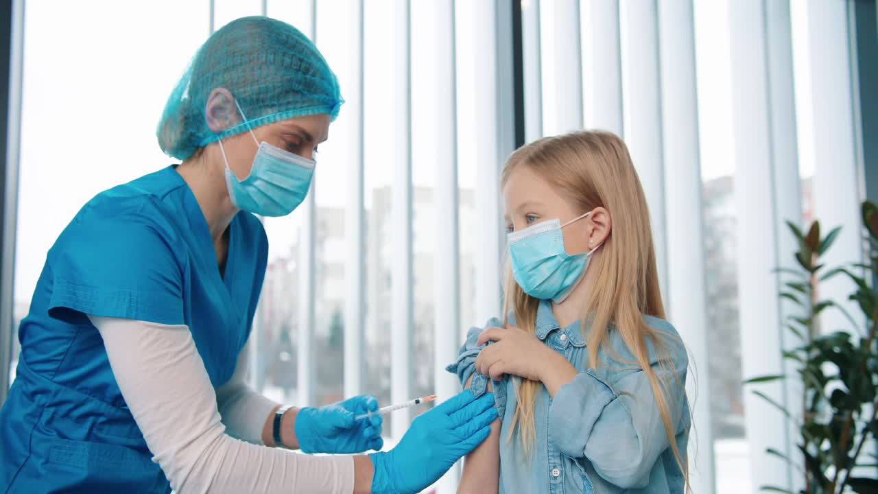 Close Up Portrait Of Cute Little Girl Child Receiving Covid 19 Vaccine In Hospital, Woman Doctor Or Nurse In Medical Mask Injecting Coronavirus Vaccine To Small Cute Kid In Medical Mask