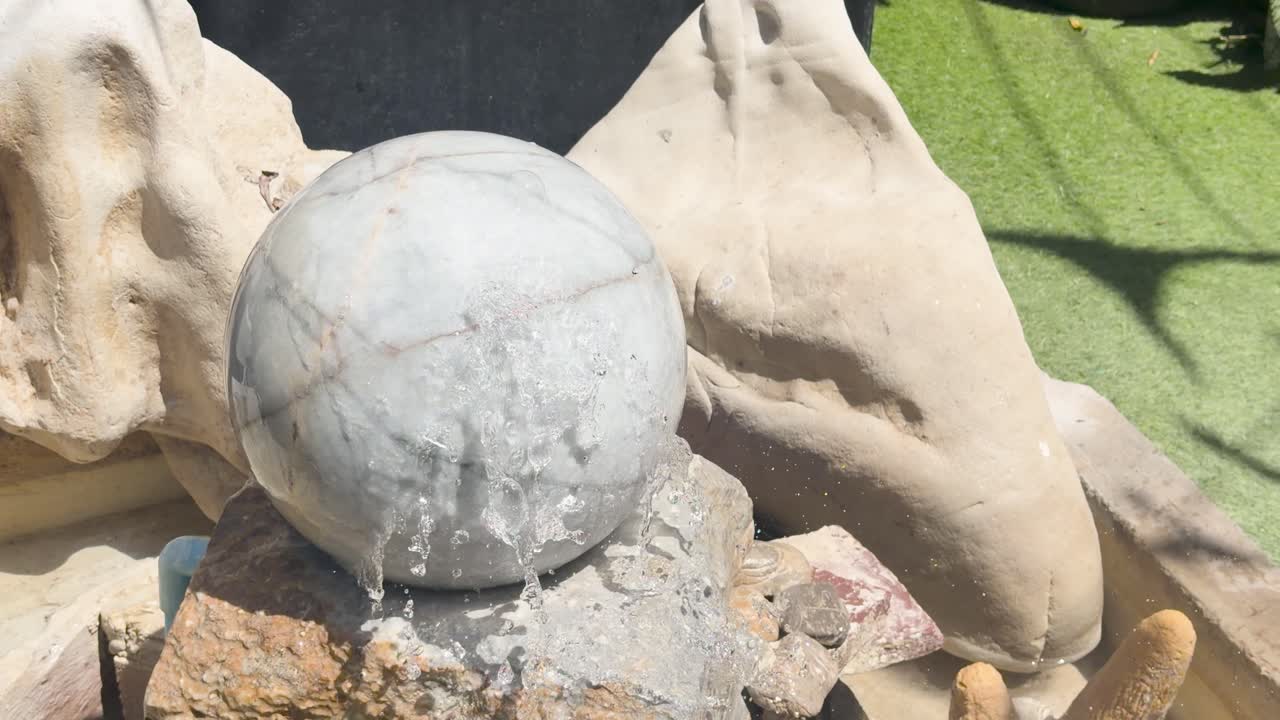 A close-up shot captures a spinning marble sphere fountain amidst decorative rocks and bright sunlight