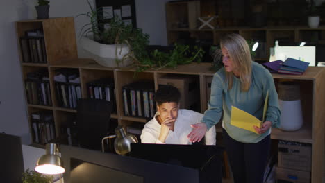 Caucasian-woman-and-biracial-man-collaborate-in-a-business-office-setting,-working-late