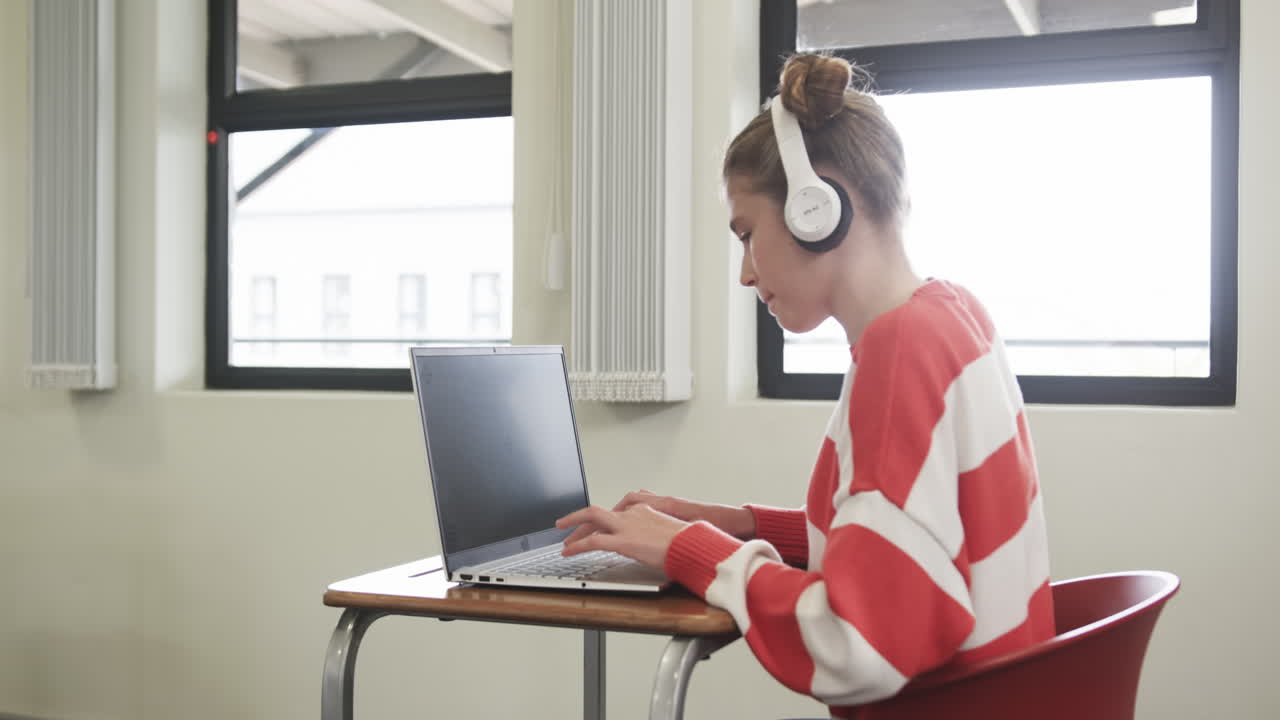 Young girl wearing headphones writing notes on laptop in classroom, focused, at school, copy space