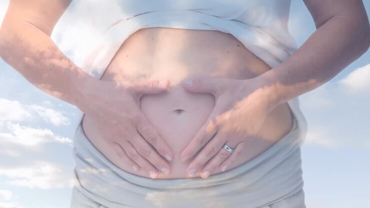 Mid section of caucasian pregnant woman rubbing her tummy against clouds in blue sky