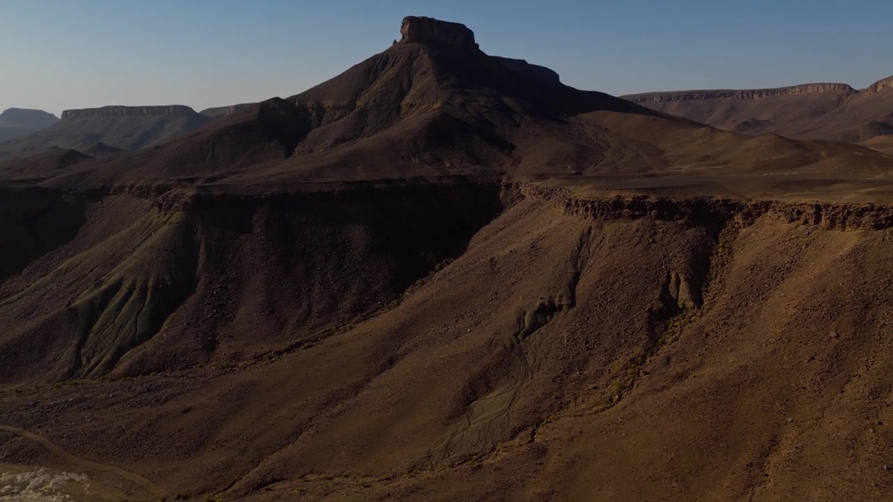 Una vista aerea attraversa una valle desertica rocciosa con un ruscello tortuoso poco profondo e arbusti sparsi, rivelando ripidi pendii e colline dalla cima piatta in Marocco