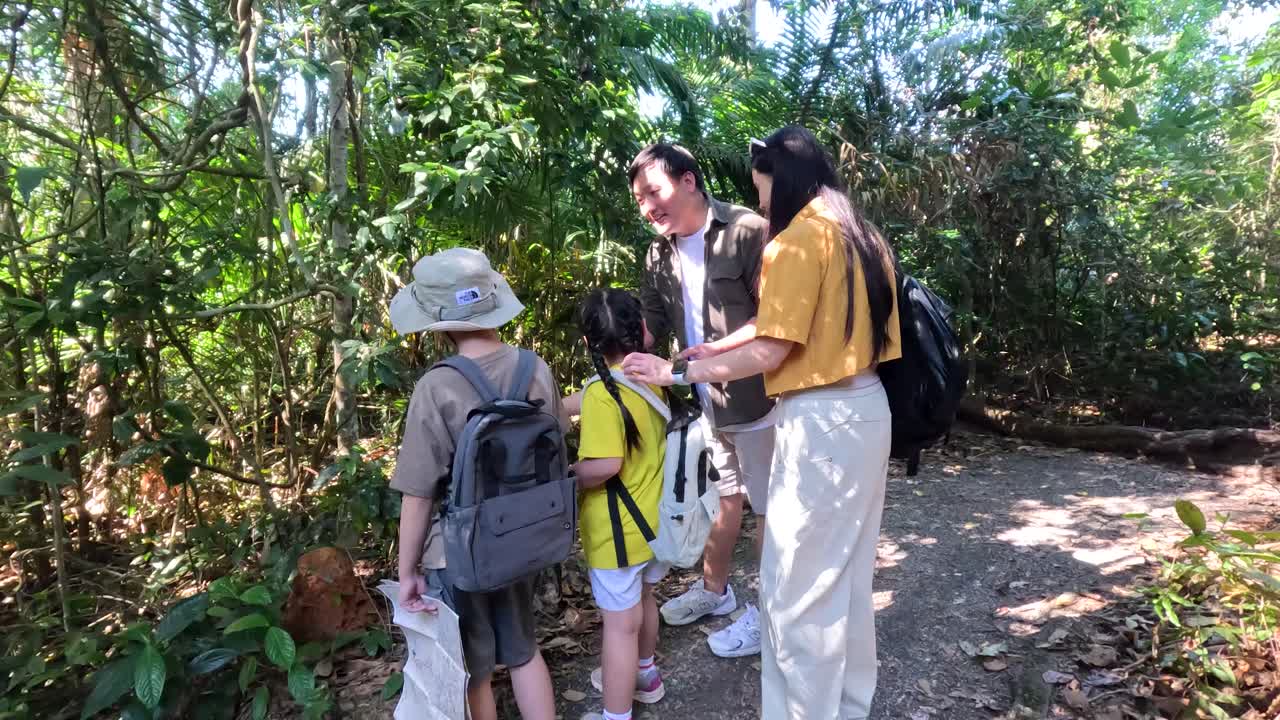 Asian family with children examines plants while hiking through a sunny tropical jungle trail