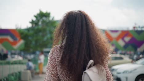 back view of lady with curly hair walking outdoors in vintage patterned blouse and white handbag, surrounded by cars and people in blurred background, colorful mural wall visible in distance