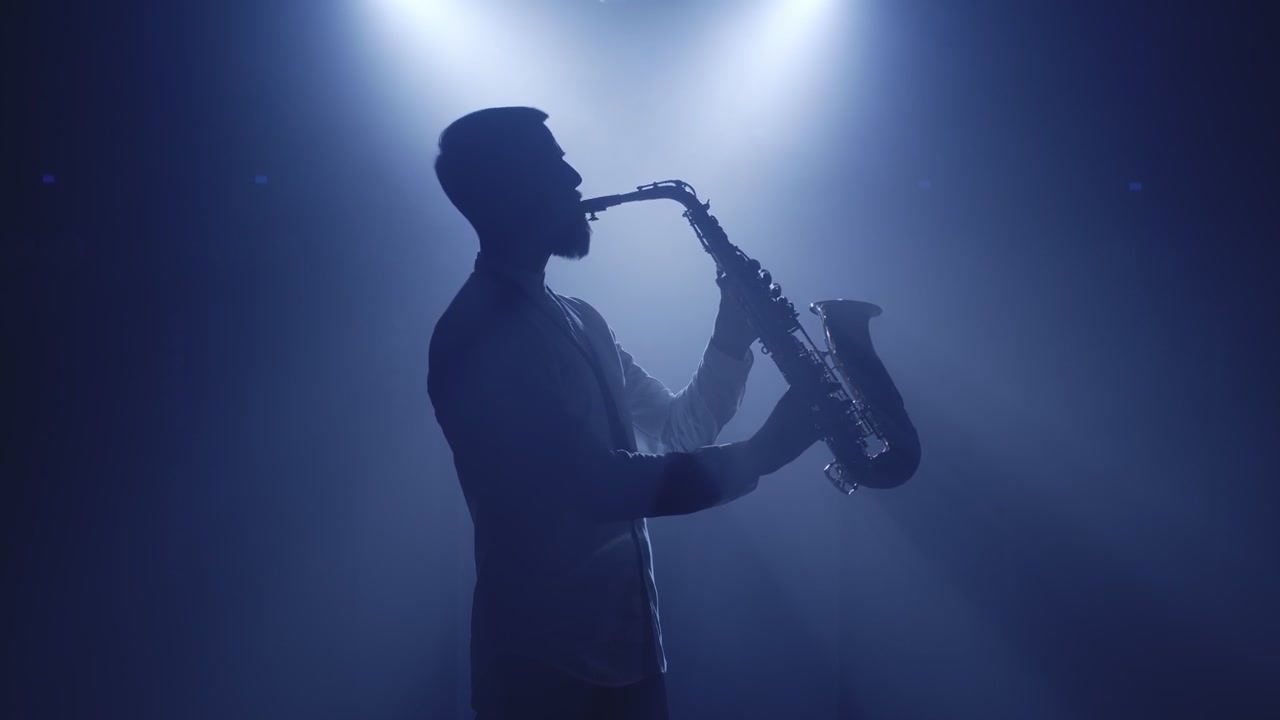 Silueta saxofonisty se saxofonem v protisvětle v temném studiu nočního klubu, který začíná hrát na saxofon.