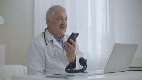 un médico está hablando por altavoz en un teléfono móvil en su oficina en la clínica grabando mensajes de voz consultando al paciente por teléfono celular