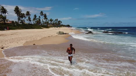 a black brazilian man in a white sunga walks toward the drone, waves, and jogs on a sunny bahia beach. the drone ascends, unveiling a breathtaking view of the coastline and ocean.
