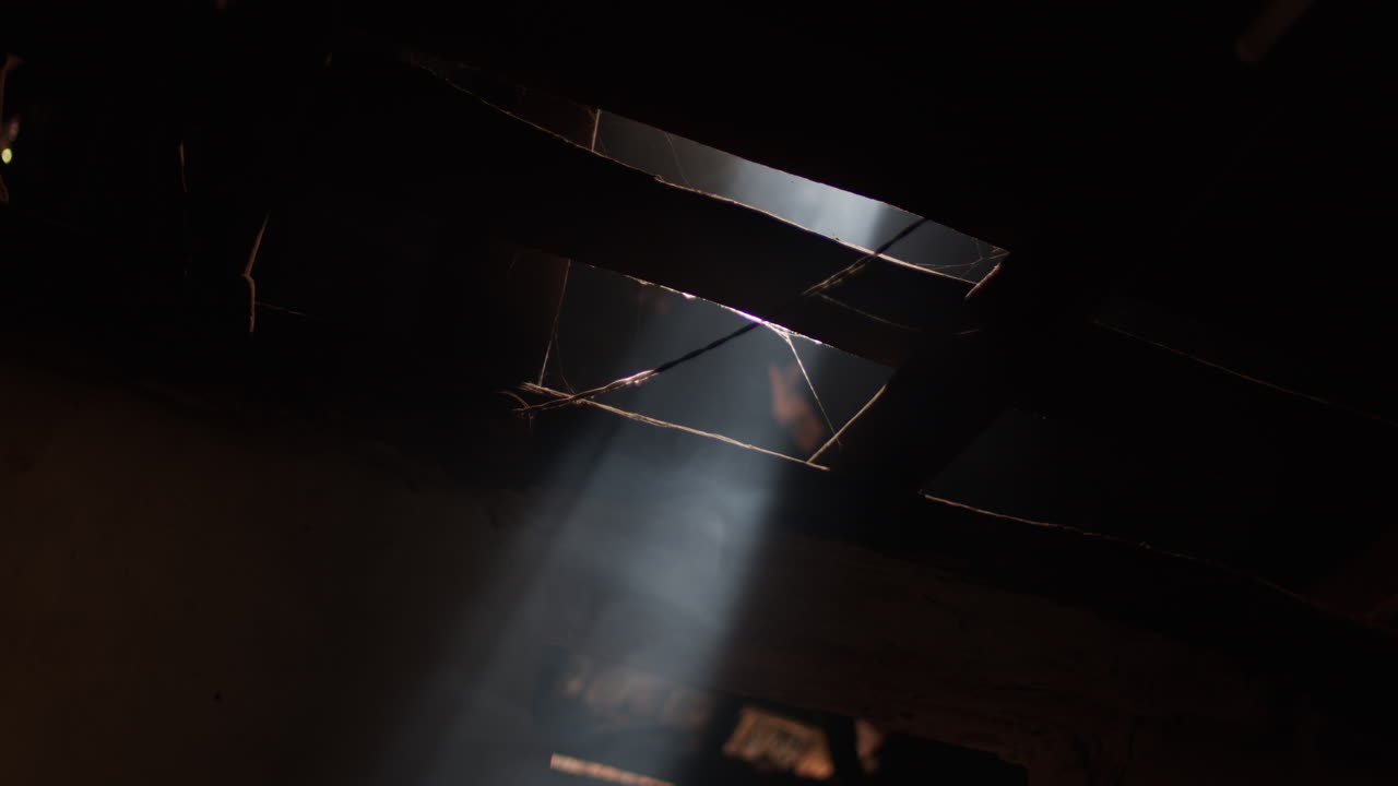 Sunbeam shines light into darkness through hole in roof of old building. Wooden rafters, wires and cobwebs shine in beam of sunlight through small opening.