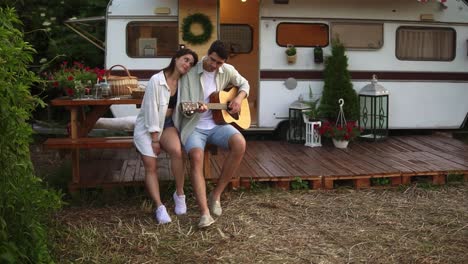 pleased, relaxful couple sitting outdoors in front their home trailer and man playing the guitar for girlfriend. outdoor. girl listening music and hugging man. slow motion. front view