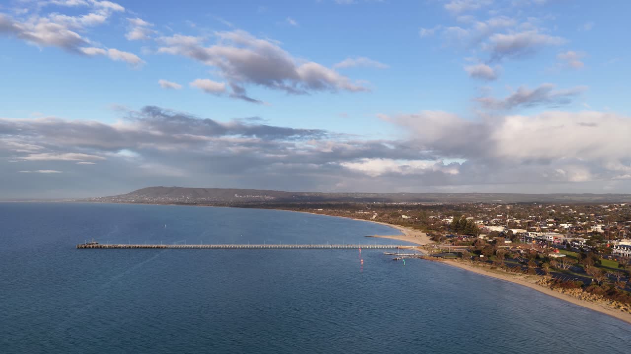 Drone pans above Rye Pier, sandy shoreline, and coastal suburb under soft evening sunlight