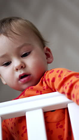 adorable baby boy standing in his cot looks at camera from top. lovely kid notices something aside, smiles adorably and hurries there. low angle view. vertical video