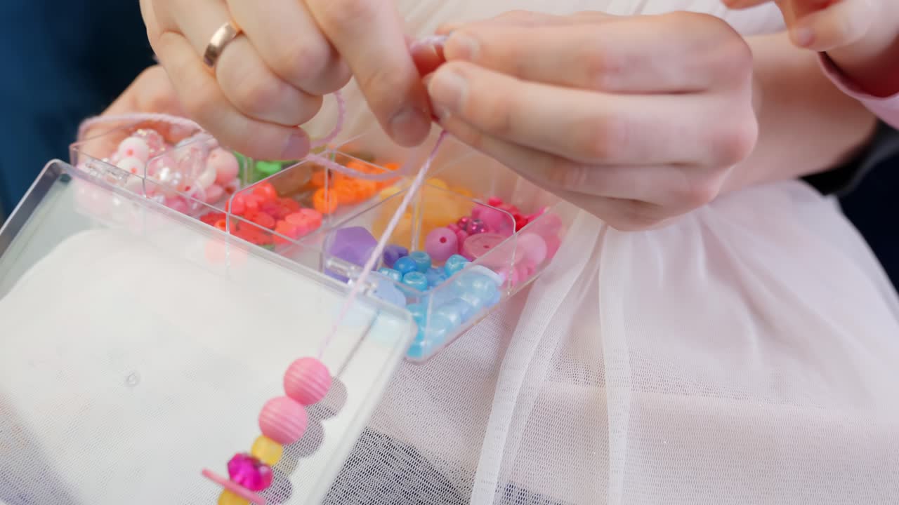 Close up view, mother's hands teach her baby to string beads for necklace with string in a soft warm environment
