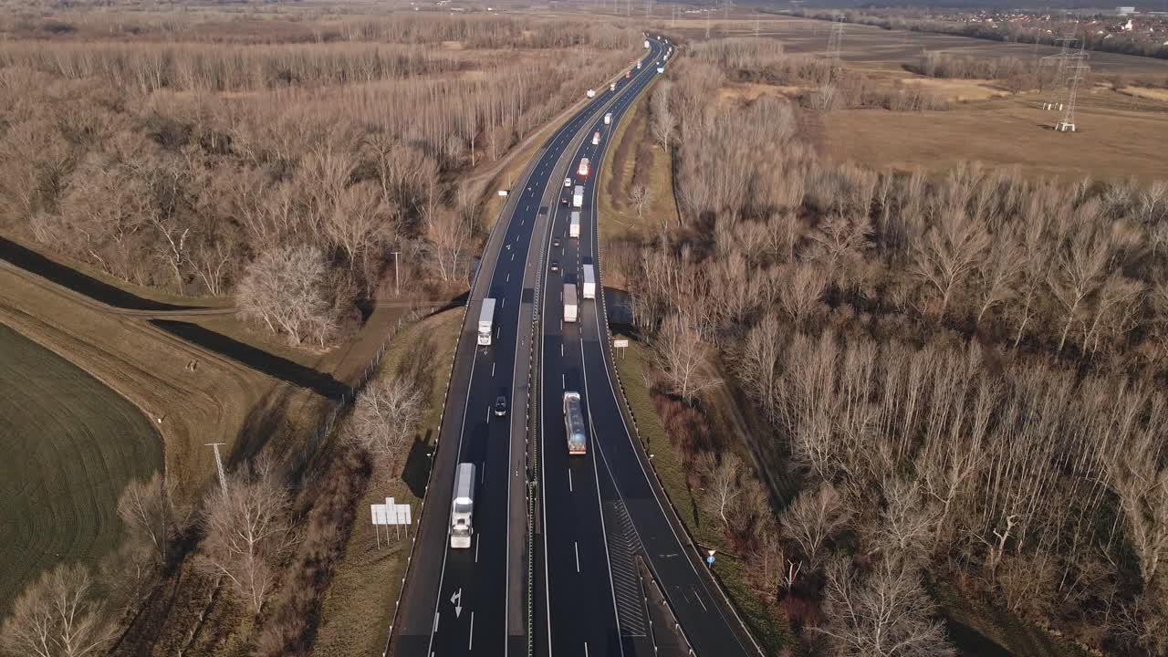 An aerial video of a highway cutting through a winter landscape with leafless trees and open fields. Several trucks and vehicles are seen traveling along the road under clear skies.