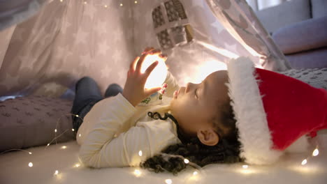 feliz chica afroamericana con sombrero de navidad sosteniendo un globo de nieve brillante en la tienda de la manta, cámara lenta