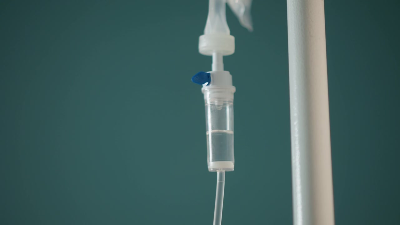 Flebo appesa a un palo color verde acqua, infermiera a domicilio regola l'infusione durante la visita, primo piano della camera di gocciolamento e dei tubi, goccioline di soluzione fisiologica visibili, morsetto e connettore a fuoco, atmosfera clinica calma ideale