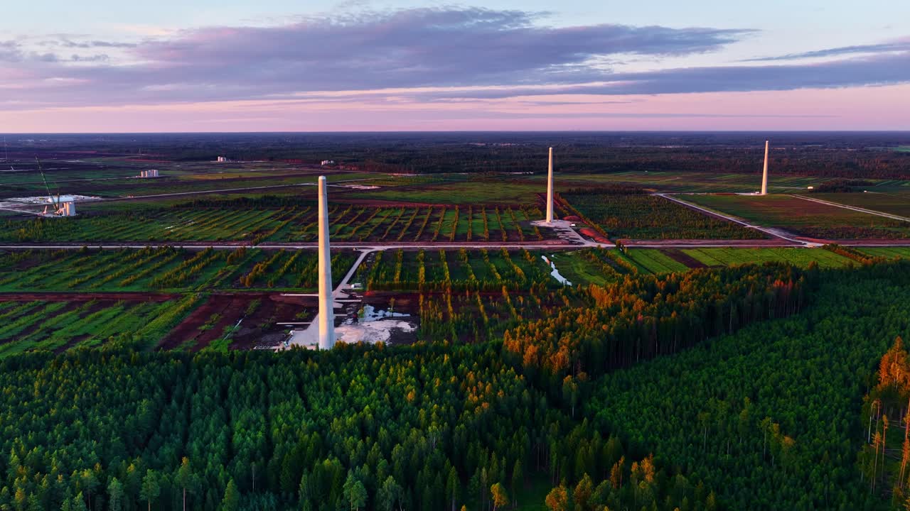 Wind turbine base being built with crane in farmland, structural parts and machinery visible across open fields by forest, aerial