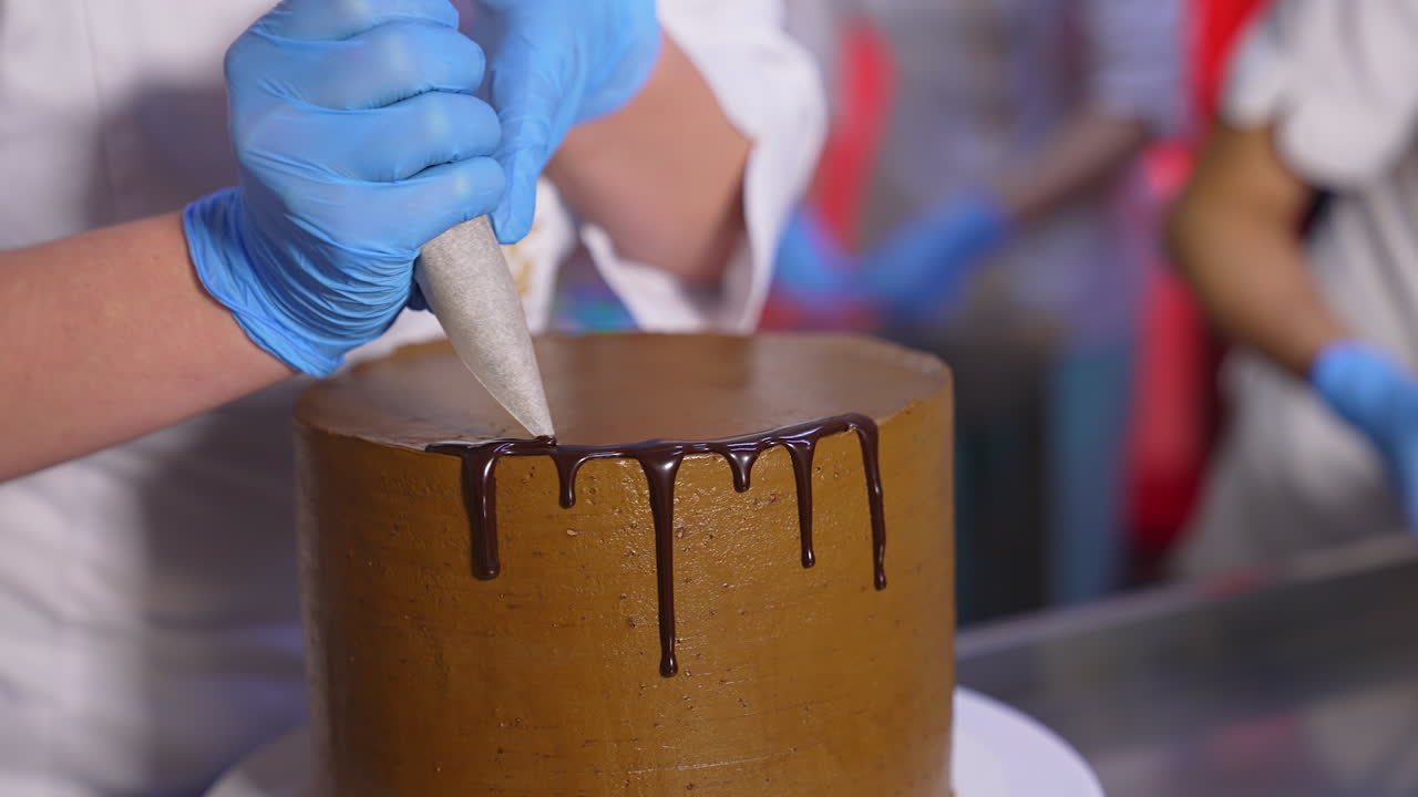 Cake decoration with chocolate. Close up. Gloved hands of confectioner squeeze liquid melted chocolate glazing on the cake edge.