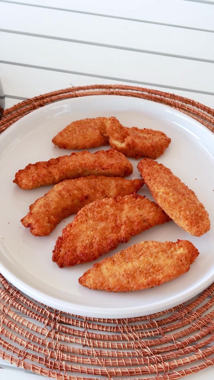 Straccetti di pollo dorati disposti su un piatto bianco con pinze che li sistemano, posizionati su una tovaglietta intrecciata