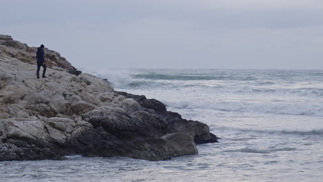 Man-walks-on-rocks-towards-wild-ocean-front,-kneels-down-and-looks-out-towards-stormy-waves-crushing-and-breaking