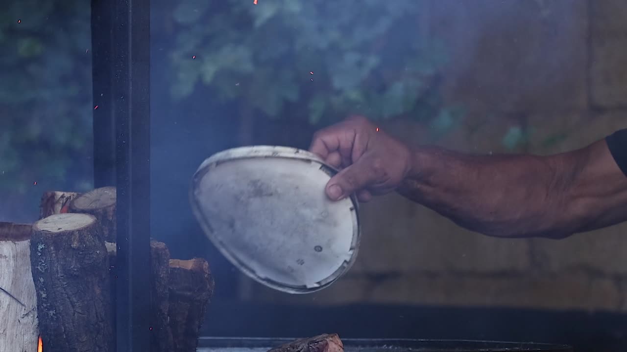 A man's arm and hand use a metal plate to fan smoke and embers outdoors