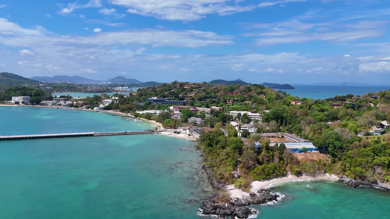 Drone footage captures Cape Panwa's lush greenery and turquoise waters under clear skies in Phuket, Thailand, showcasing serene coastal beauty