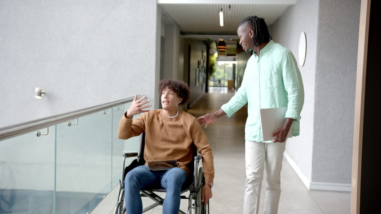 Man in wheelchair holding tablet, talking with colleague holding laptop in hallway