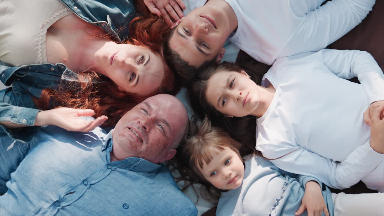 Aerial view of outdoor family lying down on picnic blanket with heads joined as older daughter talks while others listen, sharing joyful expressions, connection, and peace under soft sunlight