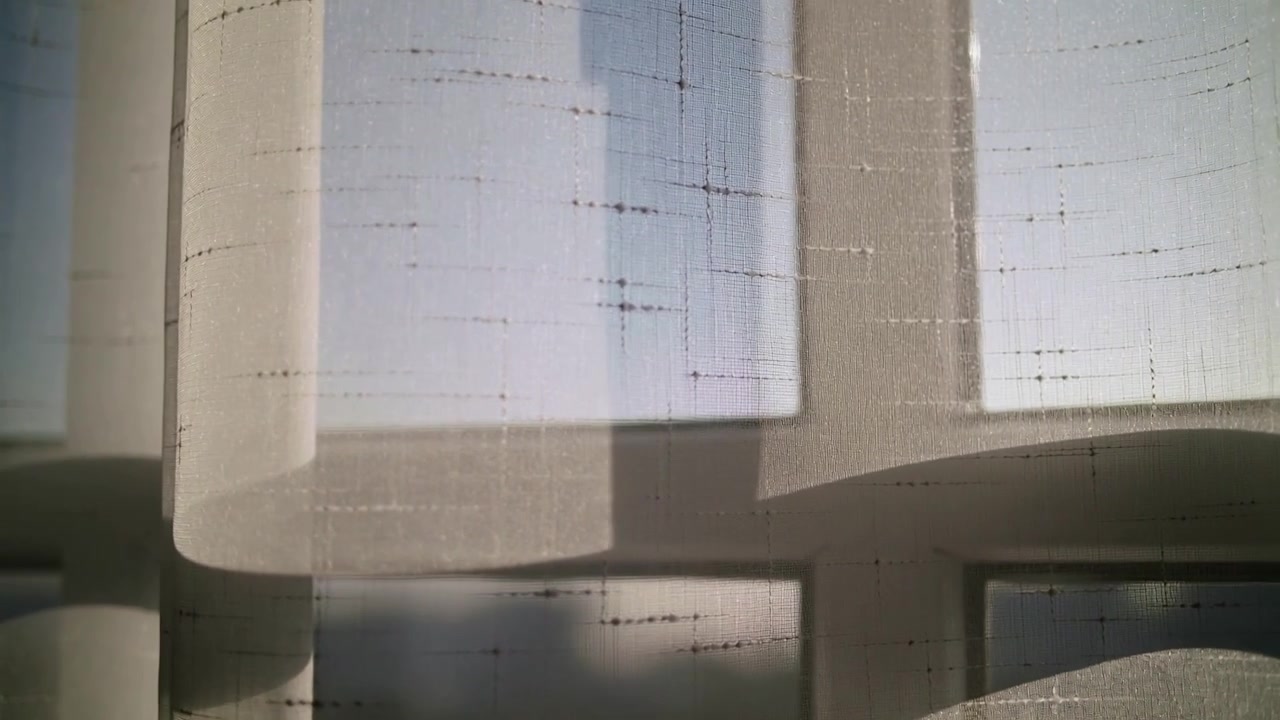 Rideau plissé transparent se déplaçant devant une fenêtre à carreaux sous l'effet d'une brise, la lumière du soleil révélant des particules de poussière.