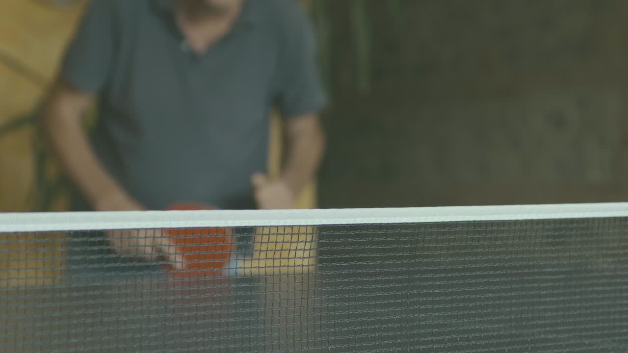 Ping pong net in the foreground and in focus, in the background not in focus a man catches the ball with a red paddle and plays with whoever is beyond the camera, slow motion, interior daytime