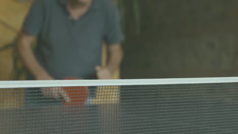 ping pong net in the foreground and in focus, in the background not in focus a man catches the ball with a red paddle and plays with whoever is beyond the camera, slow motion, interior daytime