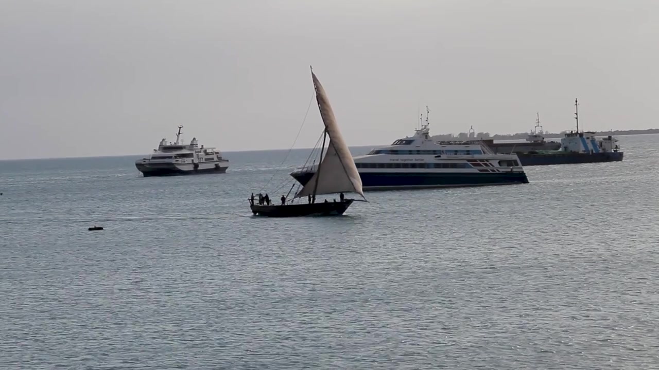 Un tradizionale dhow in legno con vela triangolare naviga in acque aperte, circondato da grandi traghetti passeggeri moderni e navi da carico in una giornata di sole.
